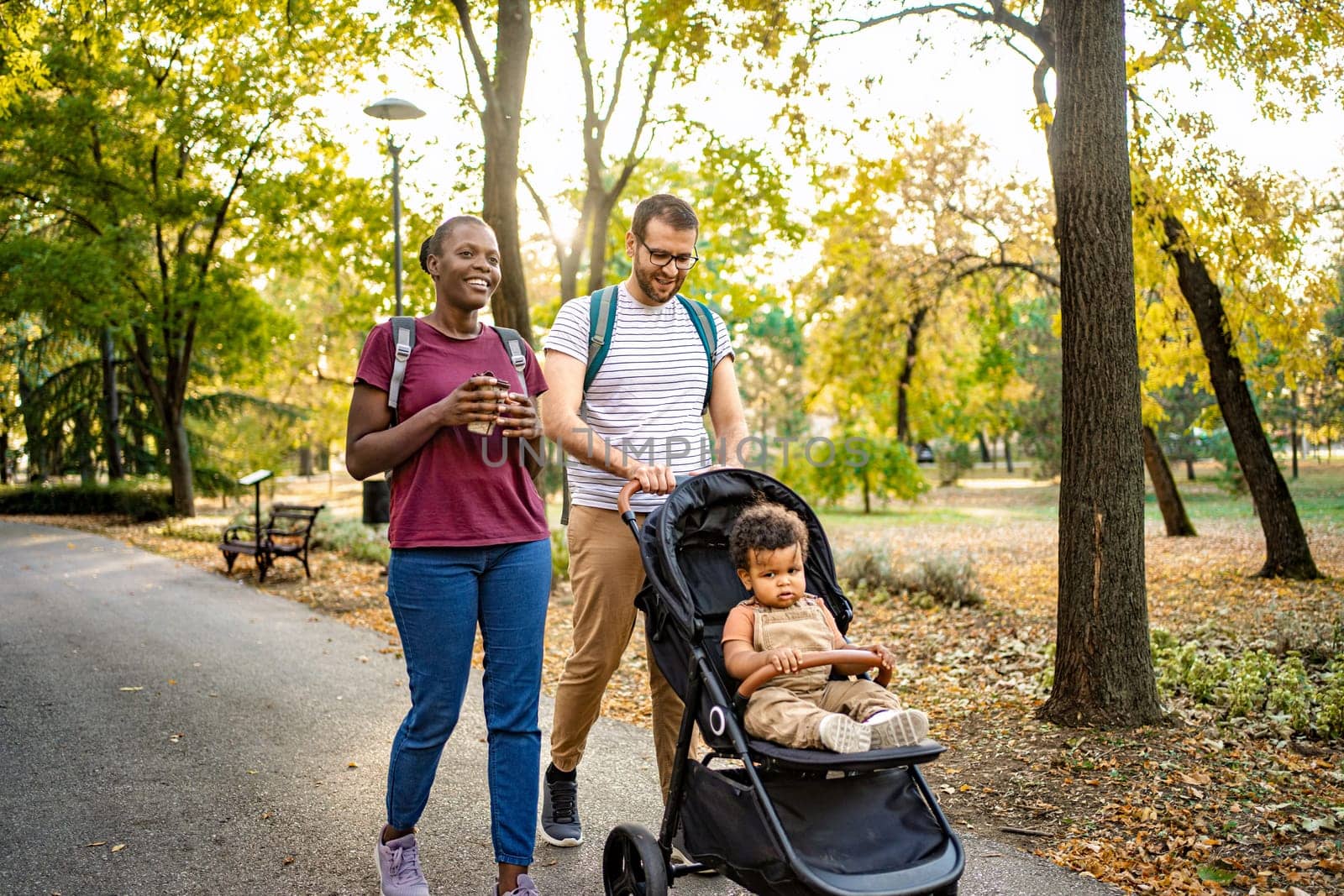 A joyful couple walks hand in hand through a picturesque park, enjoying a sunny autumn day while pushing a stroller with a curious child. Leaves create a vibrant backdrop.