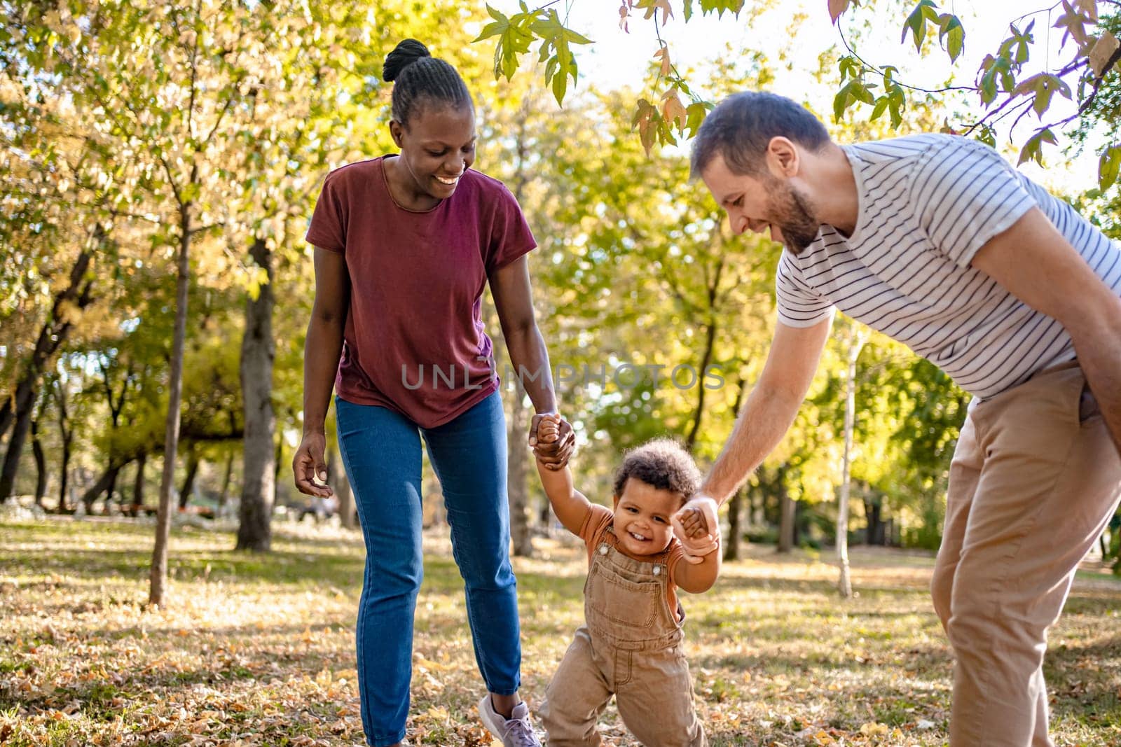 A family enjoys a sunny day in the park, with a child giggling while taking steps between two adults. The autumn foliage adds a colorful backdrop to their laughter and playful interactions.