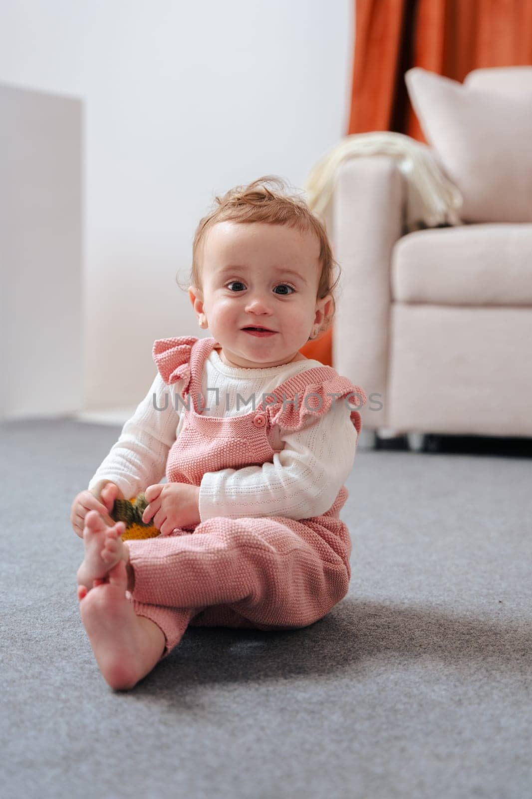 Cute baby in pink overalls sitting on a soft carpet, smiling and holding a small toy while enjoying playtime indoors in a cozy home environment with soft lighting and neutral background. Vertical