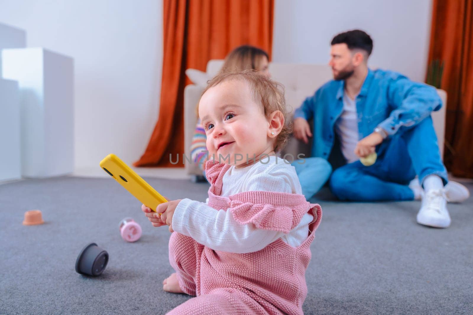 Playful baby daughter in pink overalls sits on carpet gripping a yellow smartphone, exploring technology, while her calm parents chat on the sofa in a living room,capturing daily family life at home