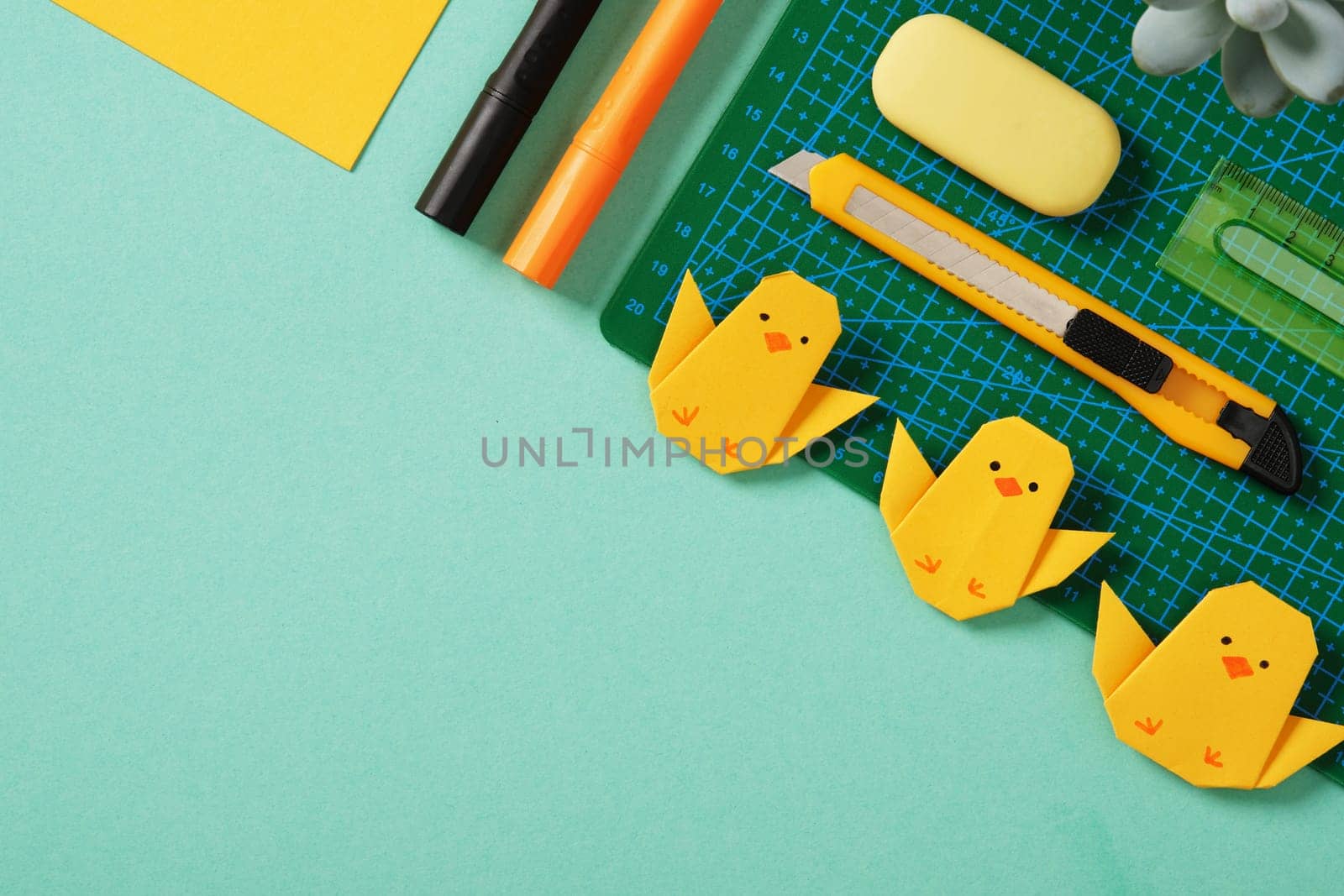Frame made of paper crafting tools: office knife, ruler, eraser and yellow paper on mint green background. Yellow origami chickens. DIY concept, copyspace
