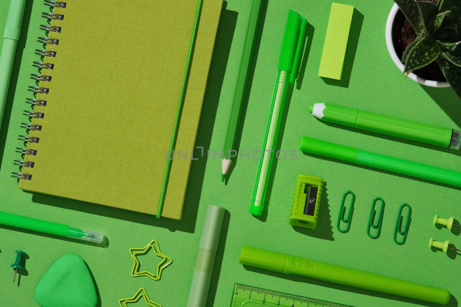 Close-up flat lay of assorted green office supplies on vibrant background, featuring spiral notebook, pens, pencil, sharpener, sticky notes, eraser, paper clips, push pins, and a small potted plant