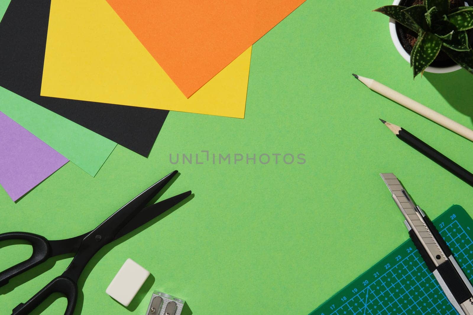 Frame of supplies for paper art hobby: scissors, craft knife, pencil, eraser, cutting mat, colored paper on green background. Copyspace.