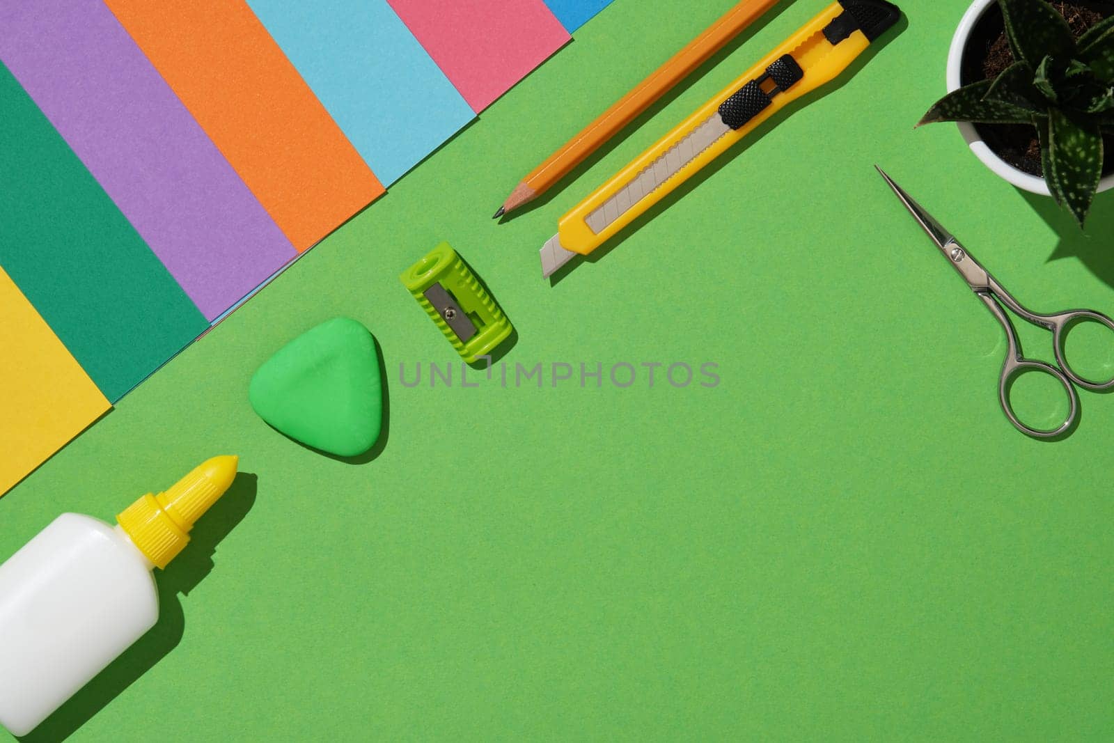 Tools for paper art hobby: scissors, craft knife, pencil, eraser, cutting mat, colored paper on green background. Copyspace. Paper crafts work table flatlay.