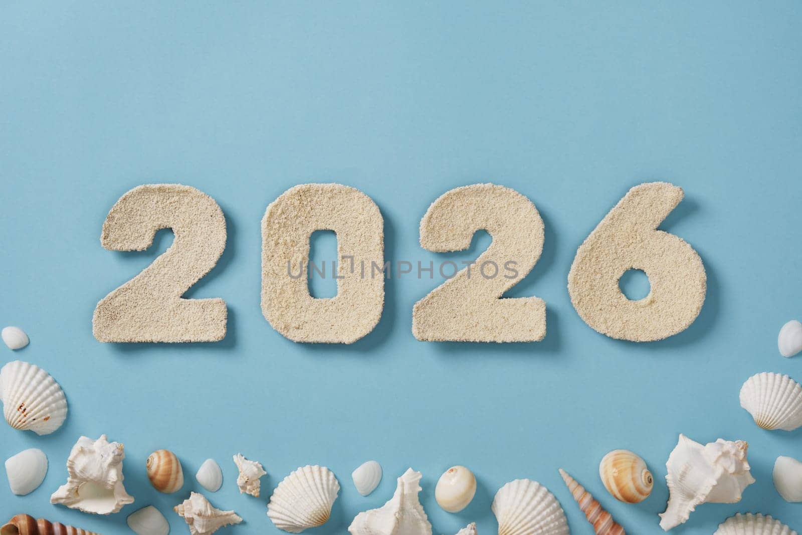 The number 2026 is crafted from fine sand and displayed against a light blue background. Surrounding the numerals are various seashells. Upcoming new year.
