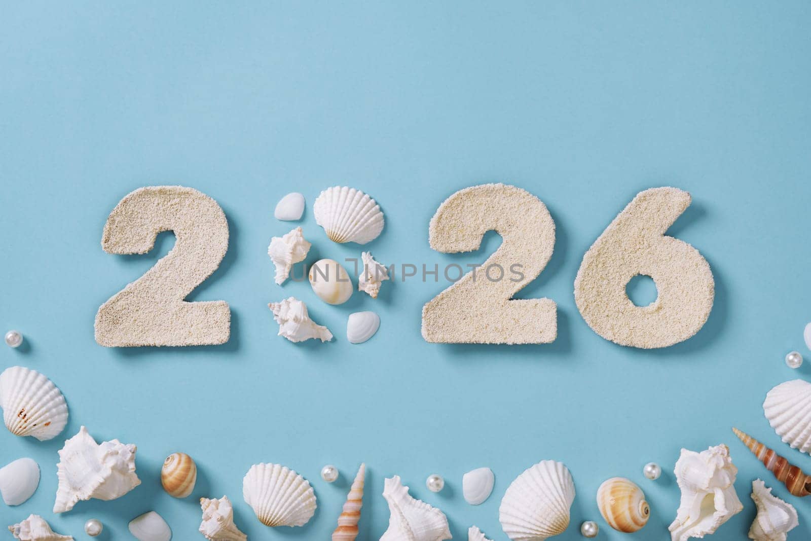 The number 2026 is crafted from fine sand and displayed against a light blue background. Surrounding the numerals are various seashells. Upcoming new year.