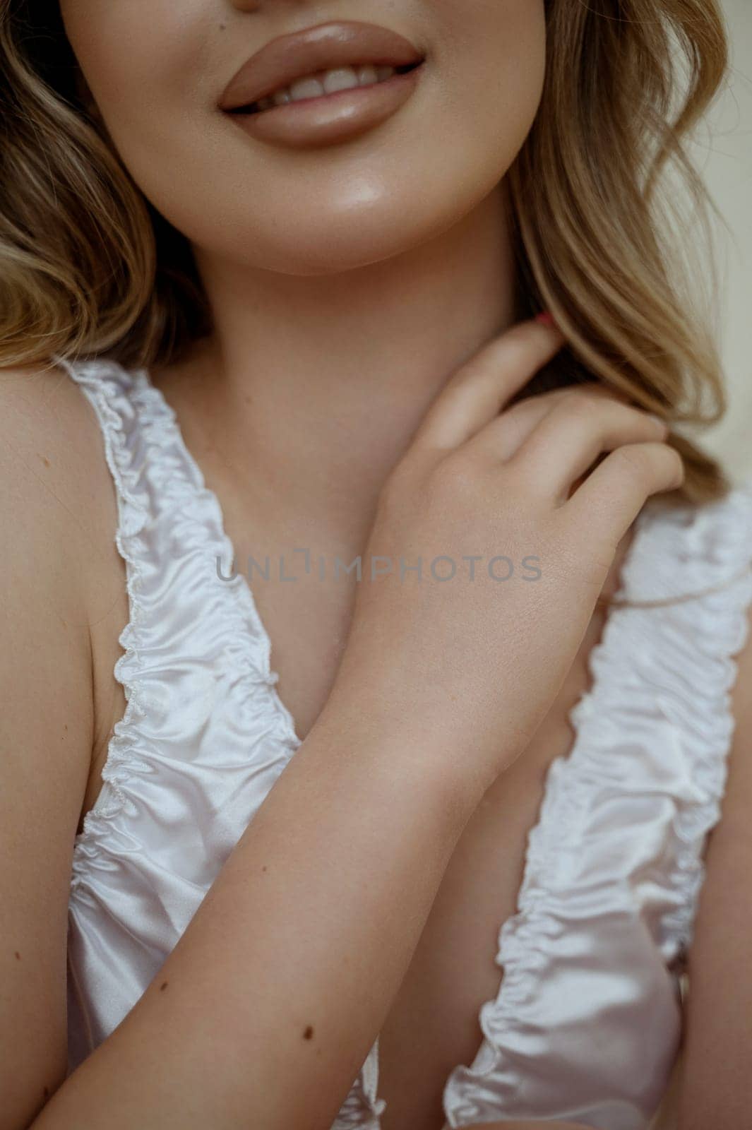 cropped close-up of attractive blonde in white silk lingerie. Lips in frame and hand. wavy hair, beautiful makeup. close-up portrait. Concept of face and skin care at home, bride's morning