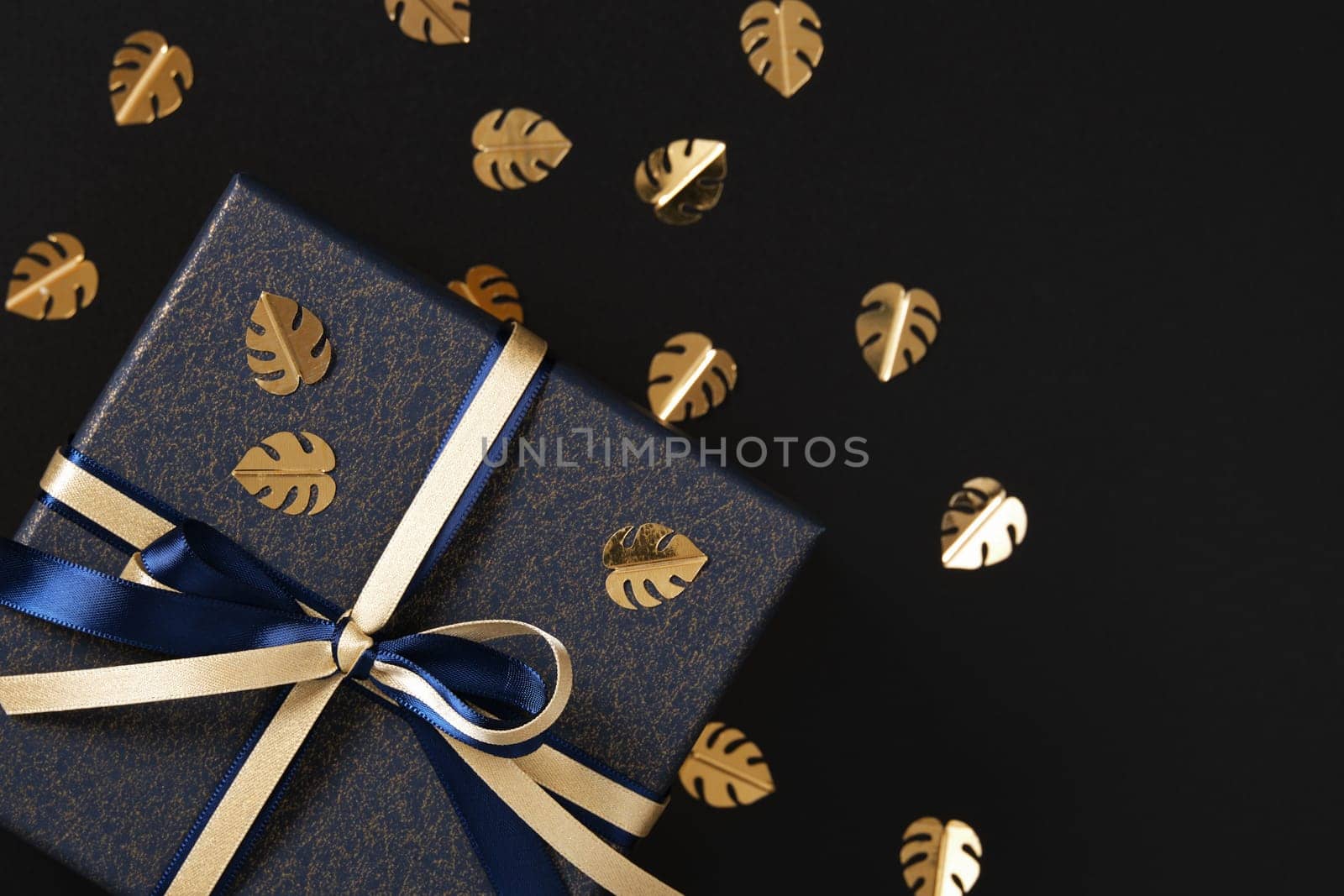 Fragment of blue gift box with gold and blue satin ribbon. Black background, gold confetti in the form of monstera leaf, space for text.