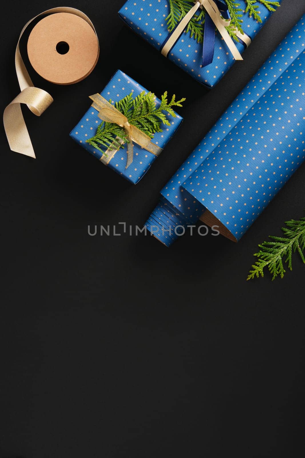 Flat composition with blue gifts with gold ribbons, brown paper tags, green thuja branches on black background. Space for your text.
