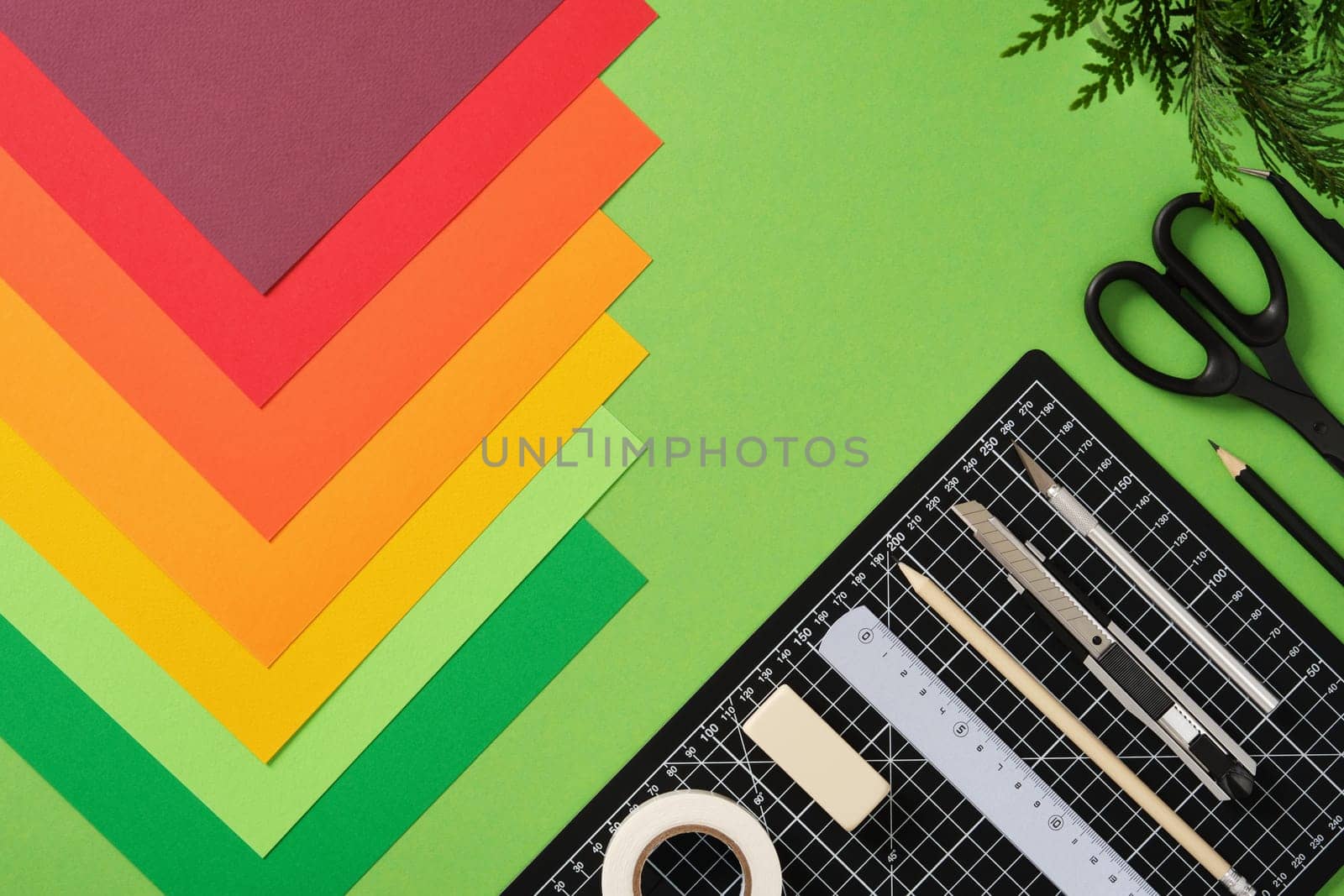 Neatly arranged paper craft supplies. Black cutting mat, cutter, scissors, ruler, pencil, tape, colored paper. Green background, space for your text.