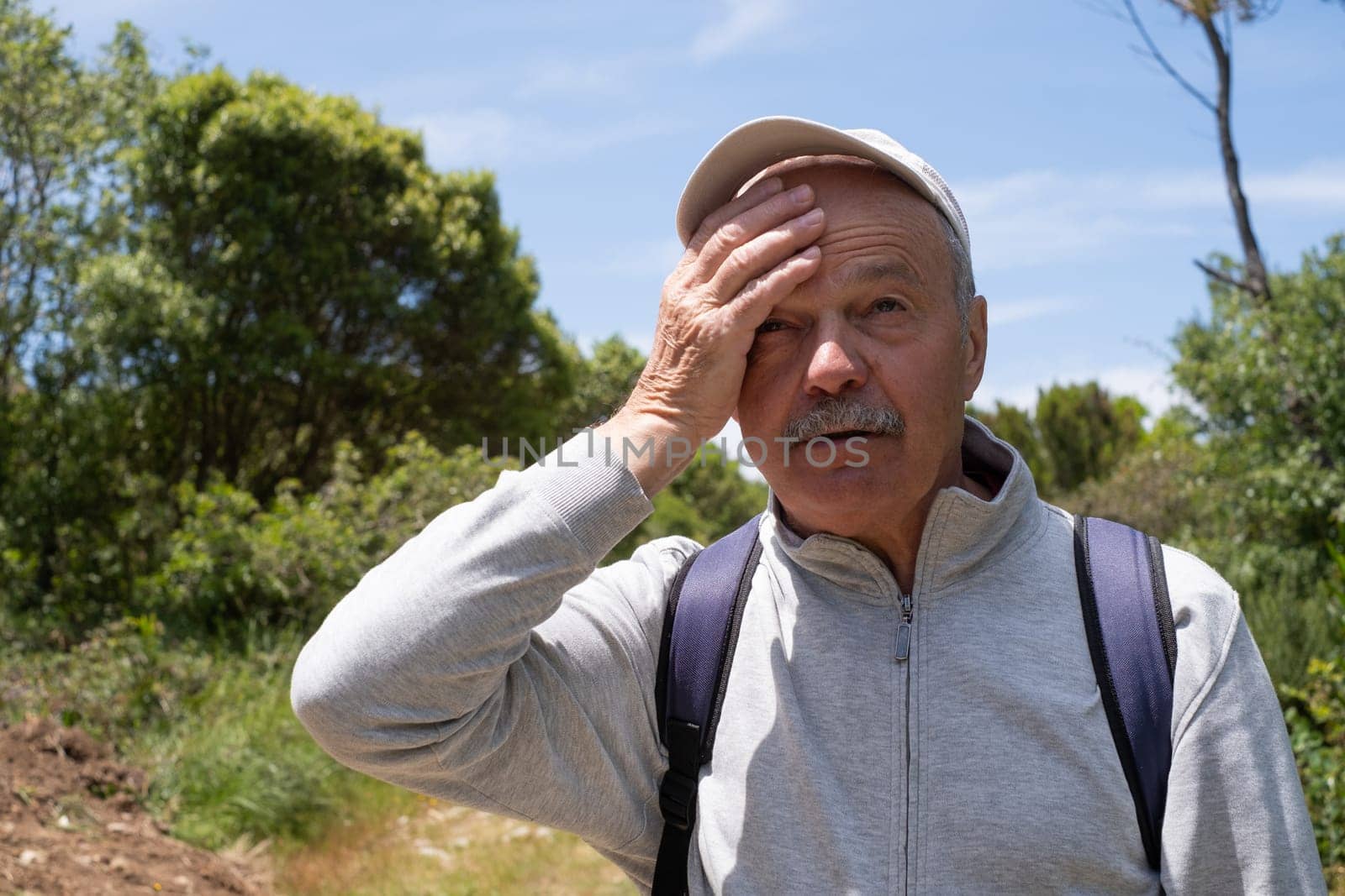 An elderly man suffering from the heat while walking in the forest, wiping sweat with his hand. High quality photo