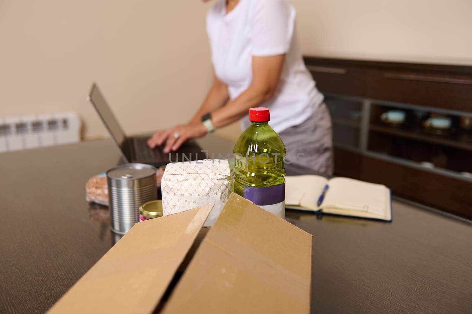 A woman is seen organizing a food package with pantry essentials like oil and canned goods while working on her laptop, suggesting donation or online shopping.