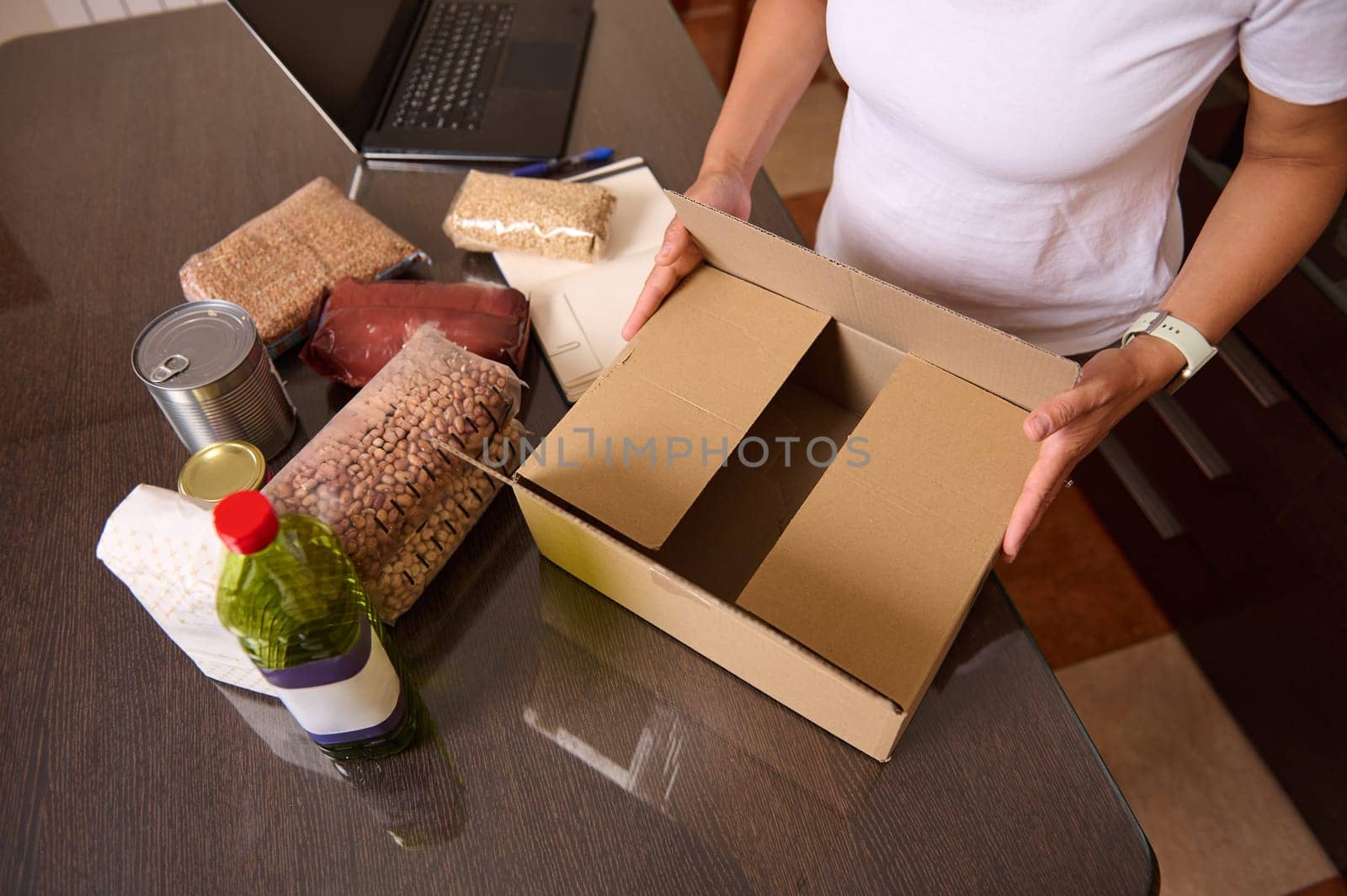 A person organizes groceries in a cardboard box, surrounded by various staple food items, displaying an act of care and preparation for storage. Ideal for charity or household themes.