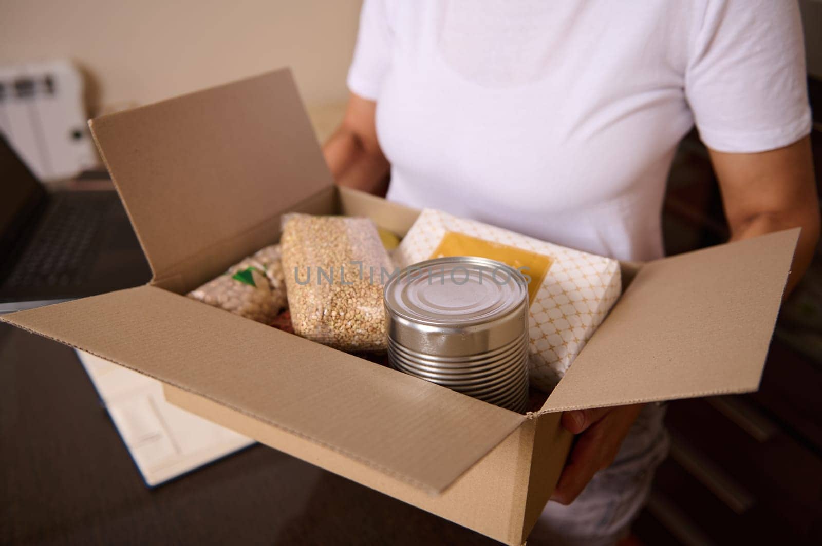 A close-up of a person holding a cardboard box with essential food supplies such as canned goods, grains, and packaging. The scene conveys charity, support, and aid distribution concepts.