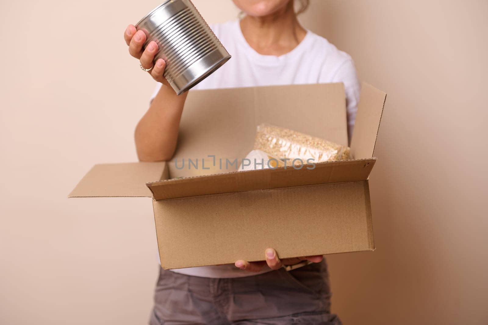 Isolated on beige studio background of volunteer hand holding a can with conserve food and packing goods into mockup cardboard box, preparing humanitarian help for charity and donation. Food Bank Day