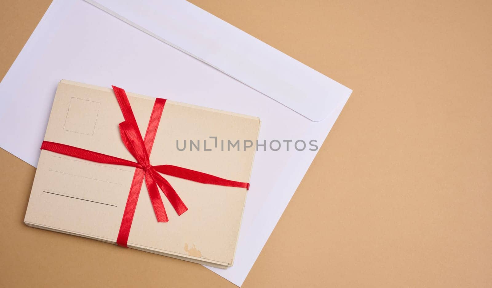 A stack of blank postcards tied with a red ribbon with a bow and an envelope lie on a beige background.
