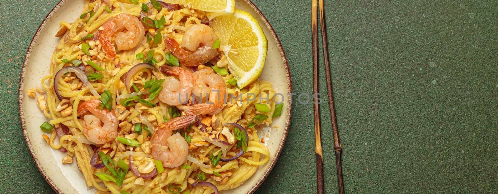 Pad thai noodles garnished with shrimp, peanuts, green onions, lemon wedges, and served with chopsticks on a rustic green background, Thai cuisine, copy space
