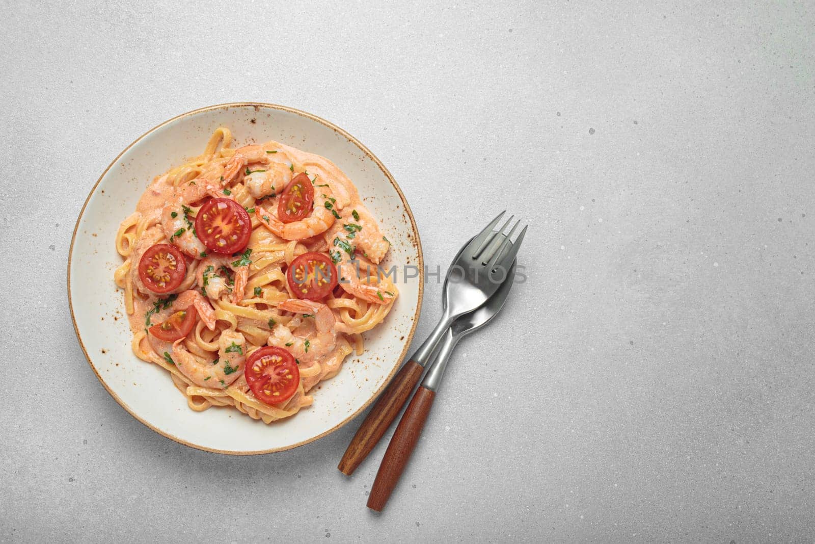 Seafood linguine pasta in creamy sauce with shrimp and cherry tomatoes, garnished with fresh parsley, served on a light grey background with cutlery top view, copy space