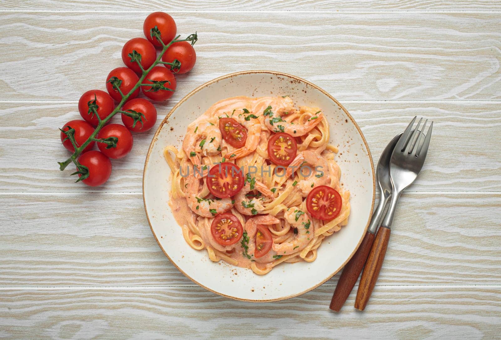 Seafood linguine pasta in creamy sauce with shrimp and cherry tomatoes, garnished with fresh parsley, served on white wooden background with cutlery top view
