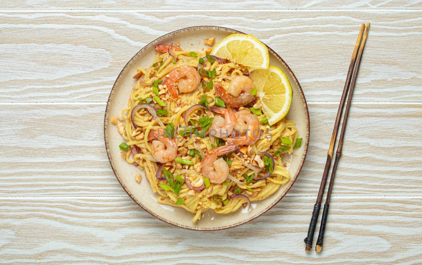 Pad thai noodles garnished with shrimp, peanuts, green onions, lemon wedges, and served with chopsticks on a white wooden table, Thai cuisine