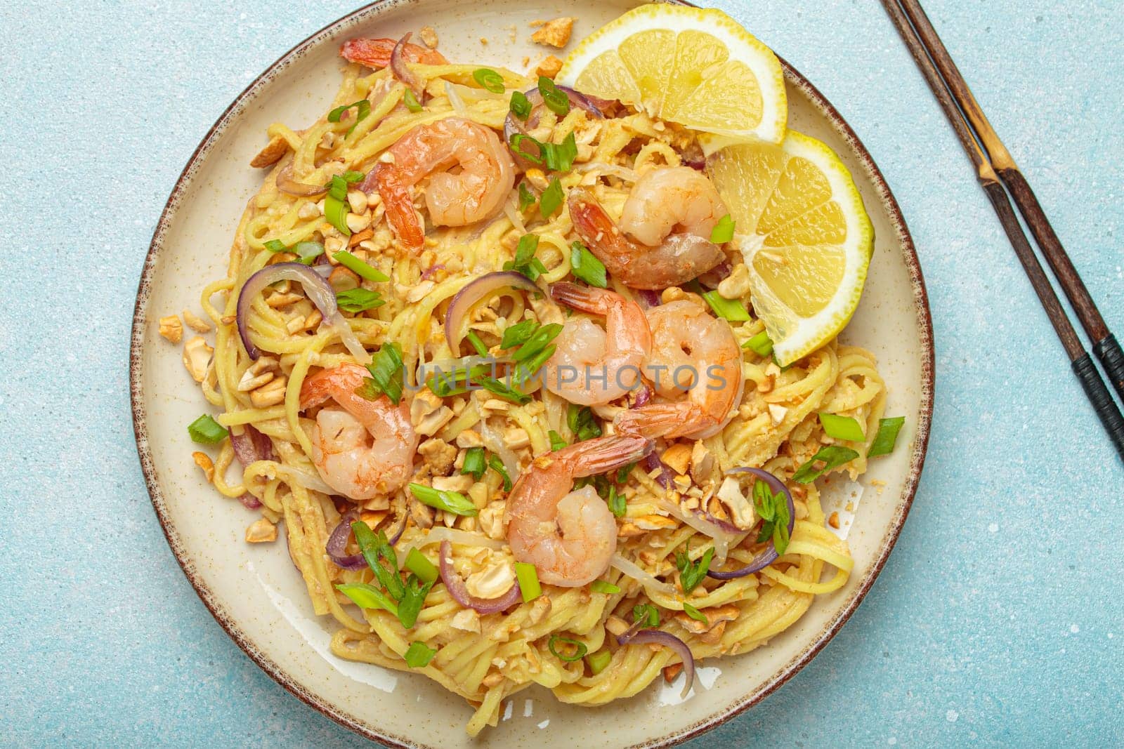 Pad thai noodles garnished with shrimp, peanuts, green onions, lemon wedges, and served with chopsticks on a light blue background, Thai cuisine