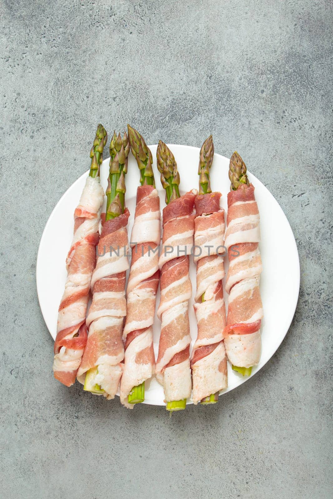 Delicious asparagus wrapped in bacon strips, seasoned with colorful peppercorns, presented on a white plate, ready to be cooked
