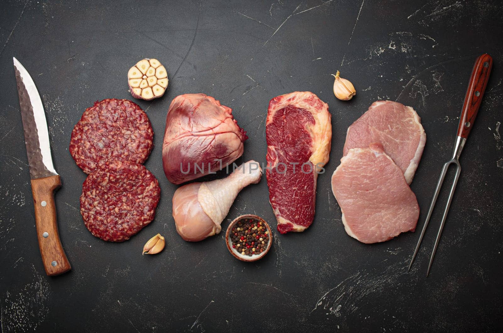 Selection of assorted raw meat cuts for zero carb carnivore diet: uncooked beef steak, pork, ground meat patty, heart, and chicken legs on black stone background top view with knife and carving fork
