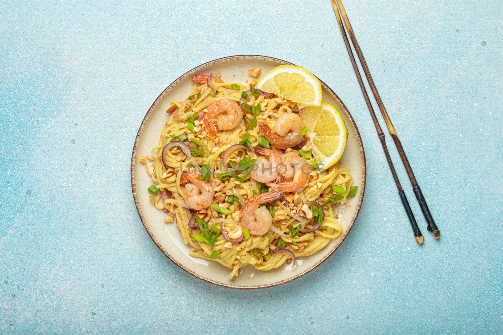 Pad thai noodles garnished with shrimp, peanuts, green onions, lemon wedges, and served with chopsticks on a light blue background, Thai cuisine