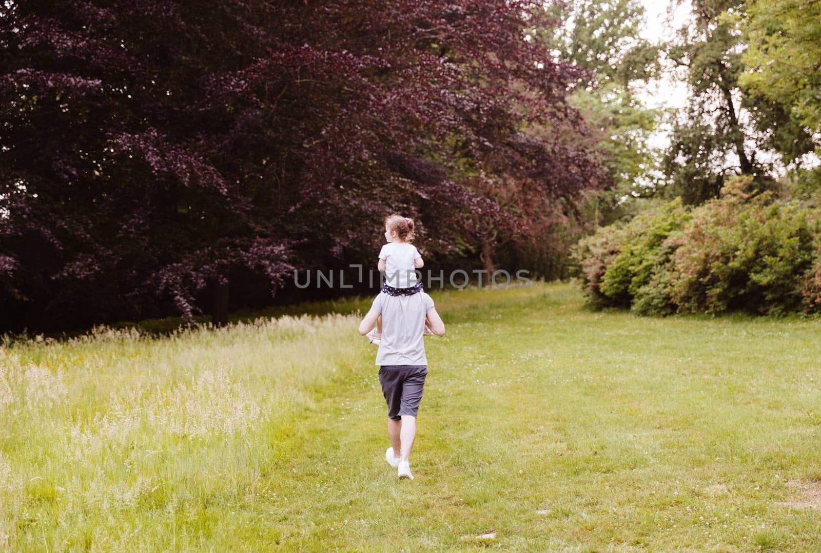 Full-length portrait from the back of a young Caucasian guy who carries a little girl on his shoulders, looking at nature around, walking in the park on a spring day, side view medium shot. Concept time walk in the fresh air.