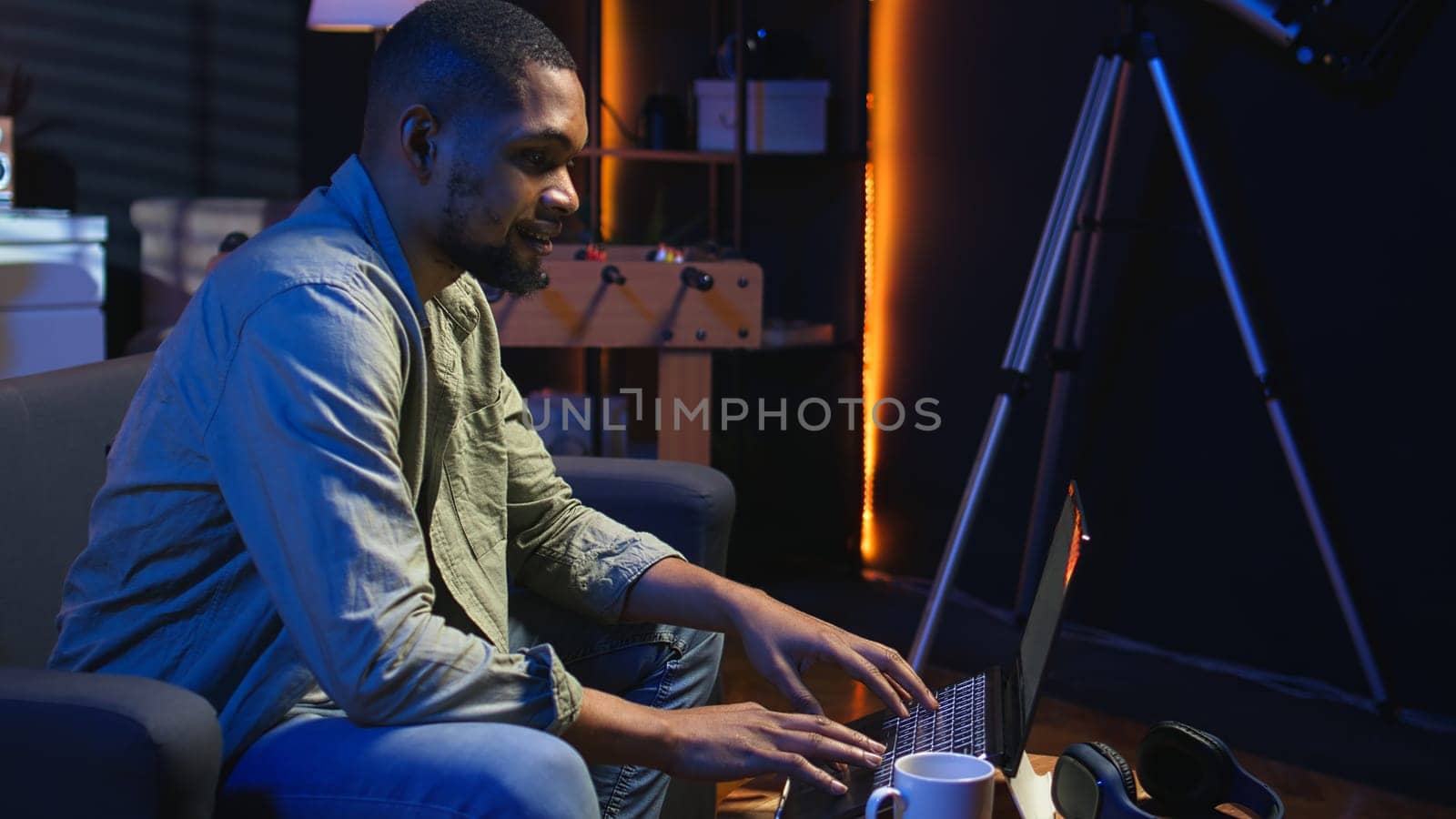 Young black adult working on his freelance tasks in the living room, adjusting his job schedule and enjoying work from his cozy home. Person navigating on web with laptop, multitasking. Camera A.
