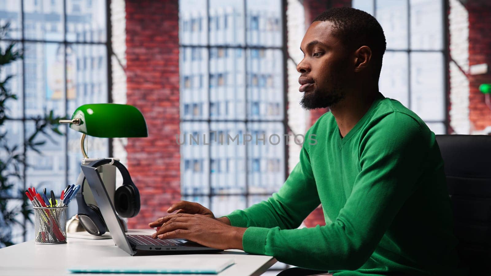 African american focused guy typing on his laptop at home office to solve daily tasks, doing remote freelance work online. Young adult surfing the web multitasking in loft apartment. Camera A.