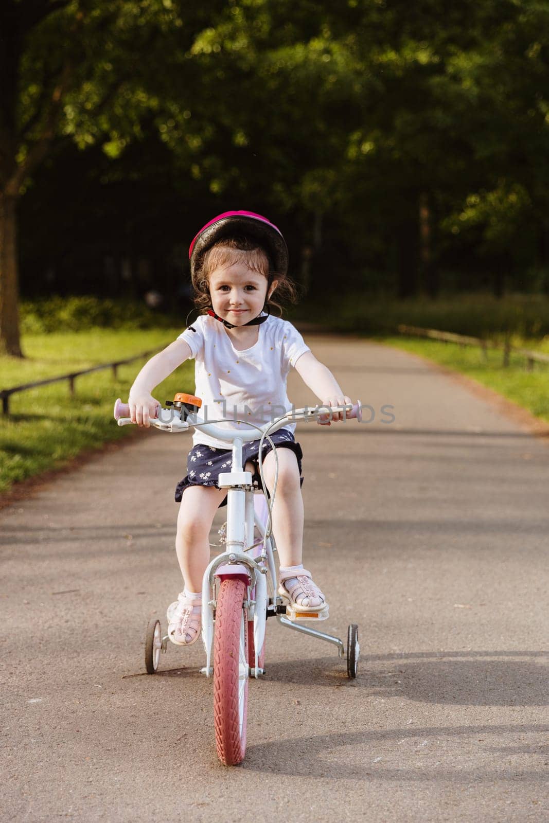 Vertical portrait of beautiful little Caucasian happy girl in bright pink helmet riding bicycle alone on path in park on spring day, bottom side view close up. Happy childhood concept, kids riding bicycles.