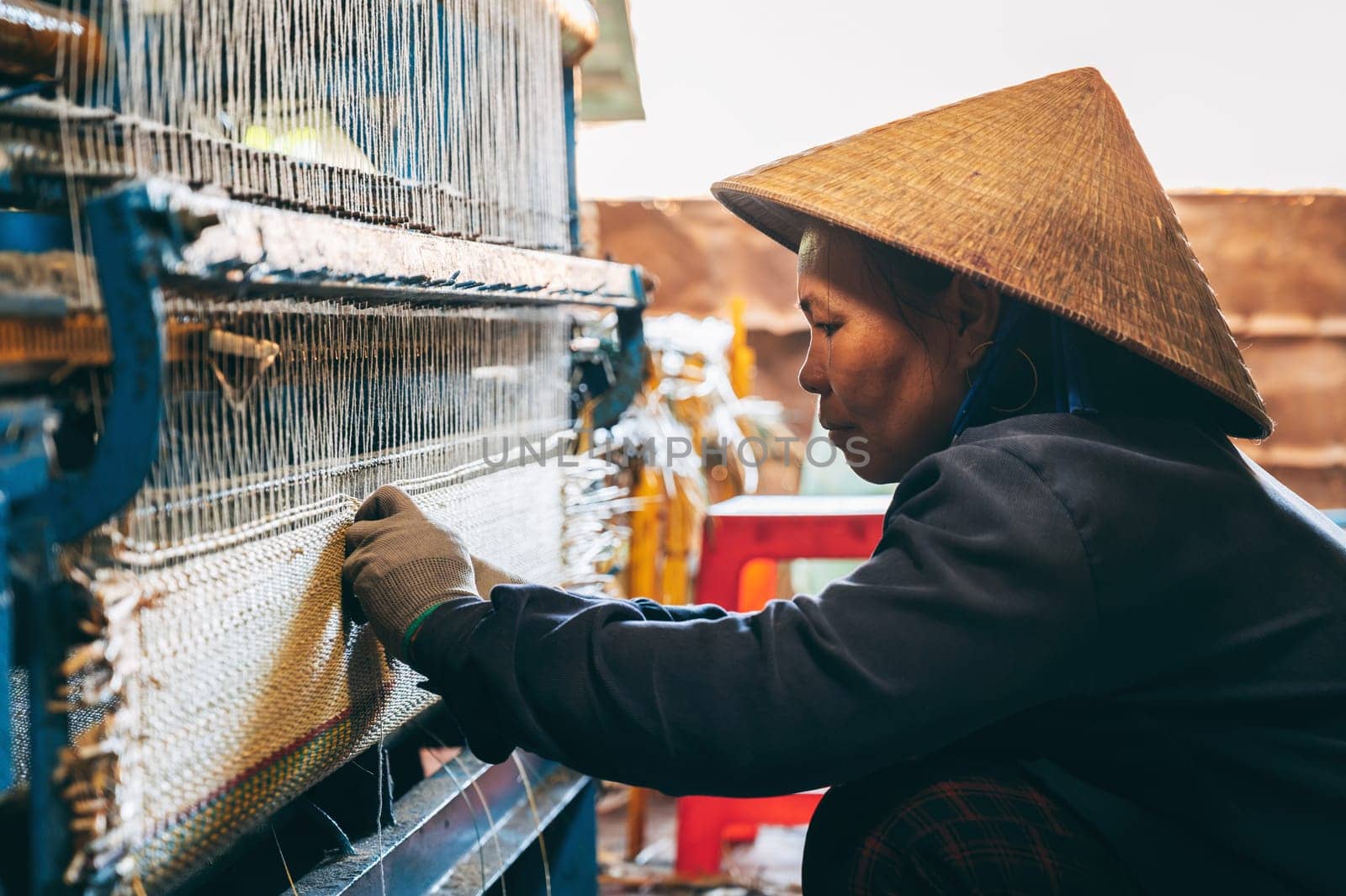 Vietnamese woman weaver in straw hat works at a loom weaving machines in workshop at factory for production of woven reed mats in Asia. An Cu, Tuy An, Phu Yen province, Vietnam - April 14, 2025