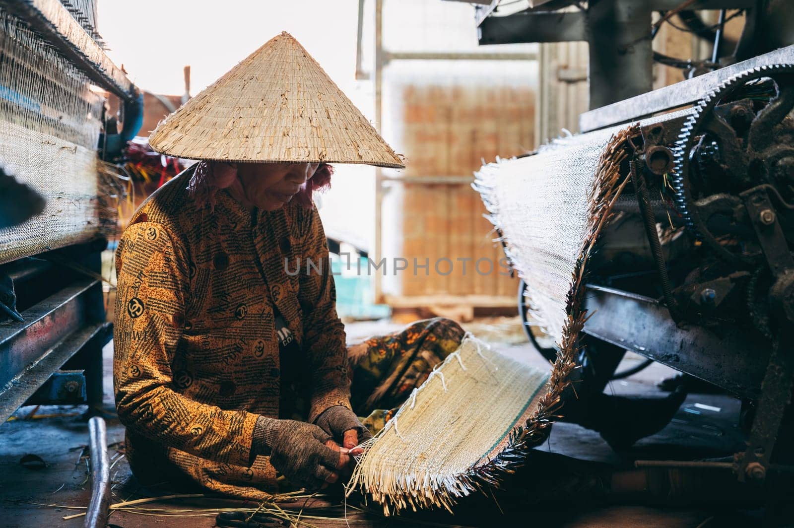 Vietnamese woman weaver in straw hat works at loom weaving machines in workshop at factory for production of woven reed mats in Vietnam. An Cu, Tuy An, Phu Yen province, Vietnam - April 14, 2025