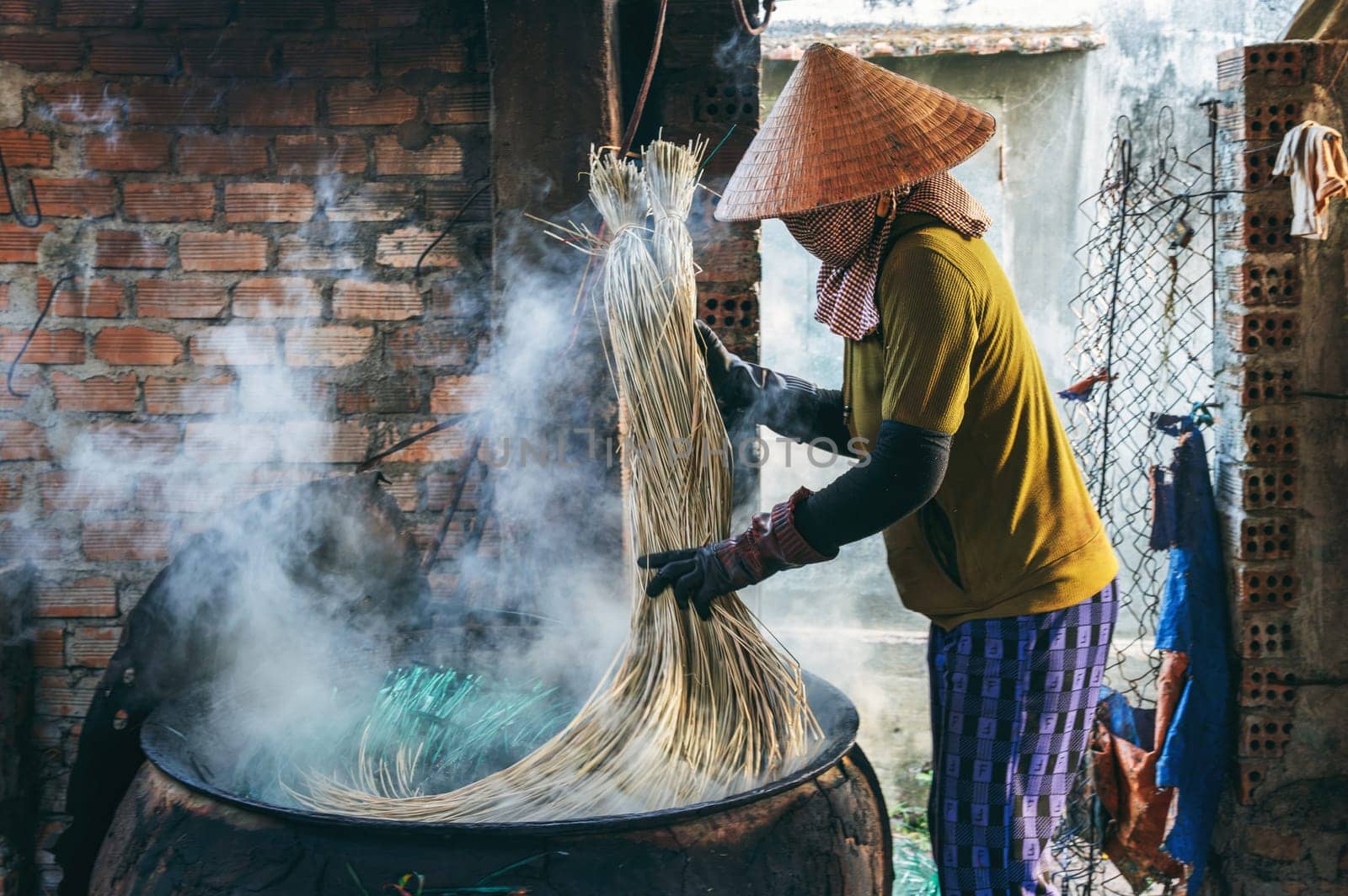 Vietnamese woman in a traditional straw hat dyes reeds at woven mat factory in Vietnam