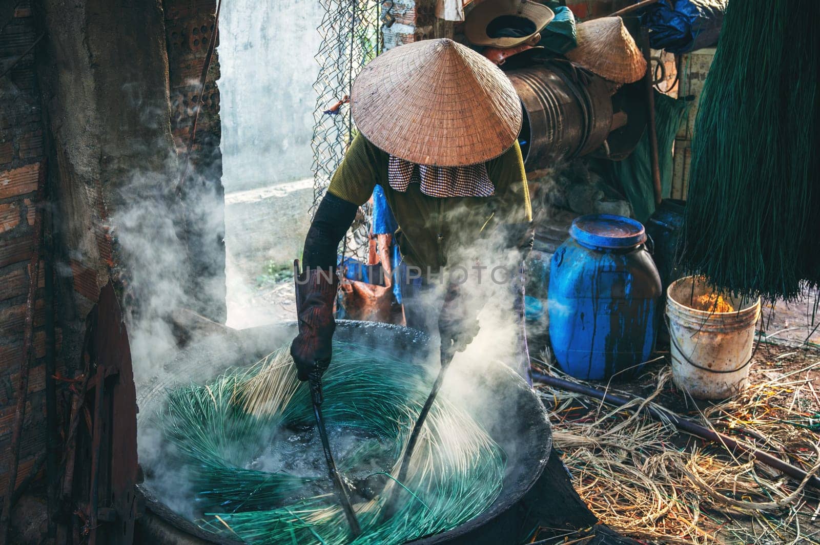 Vietnamese woman in a traditional straw hat dyes reeds at woven sleeping mat factory in Vietnam