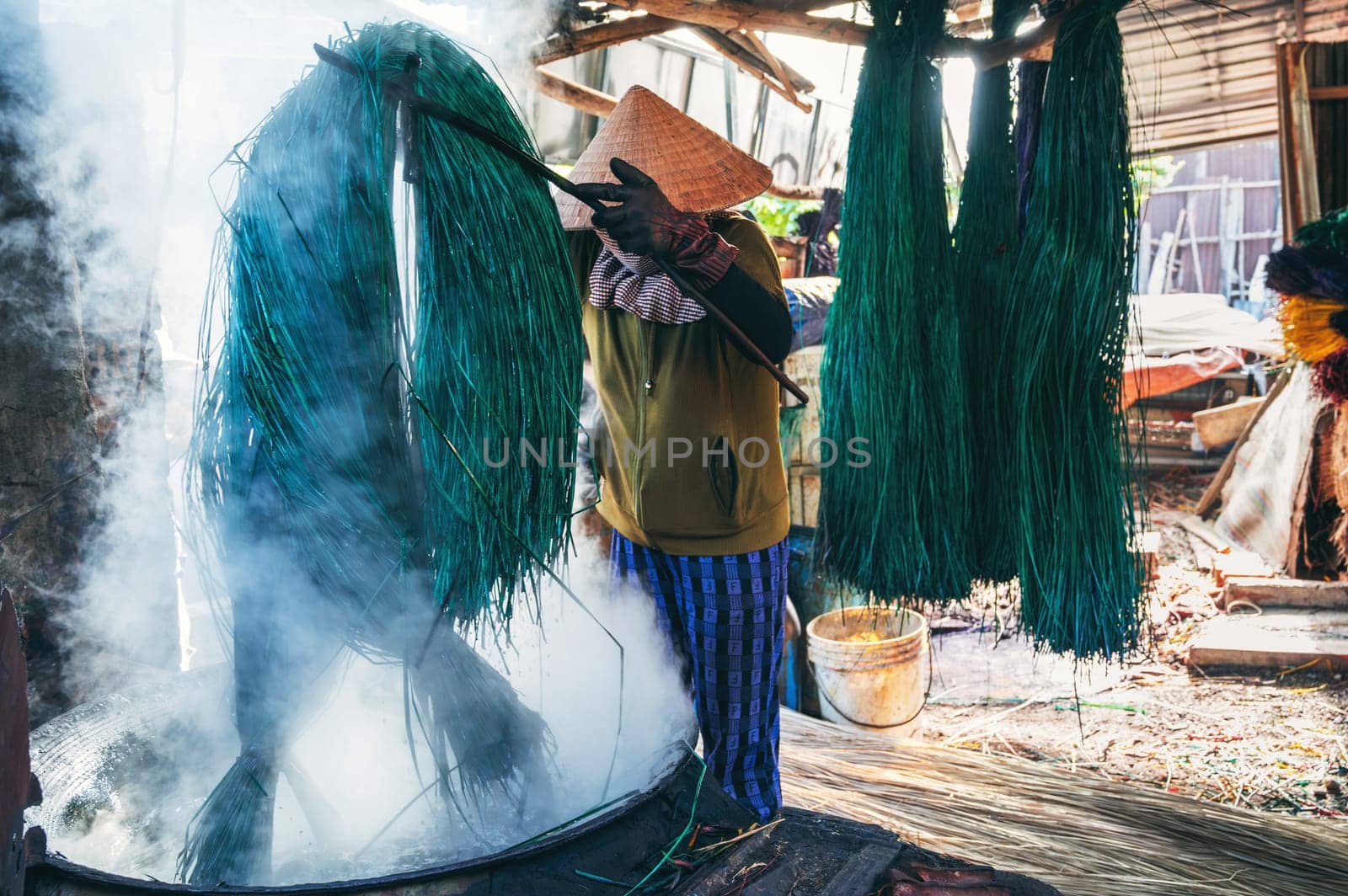 Vietnamese woman in a traditional straw hat dyes reeds at woven mat factory in Vietnam