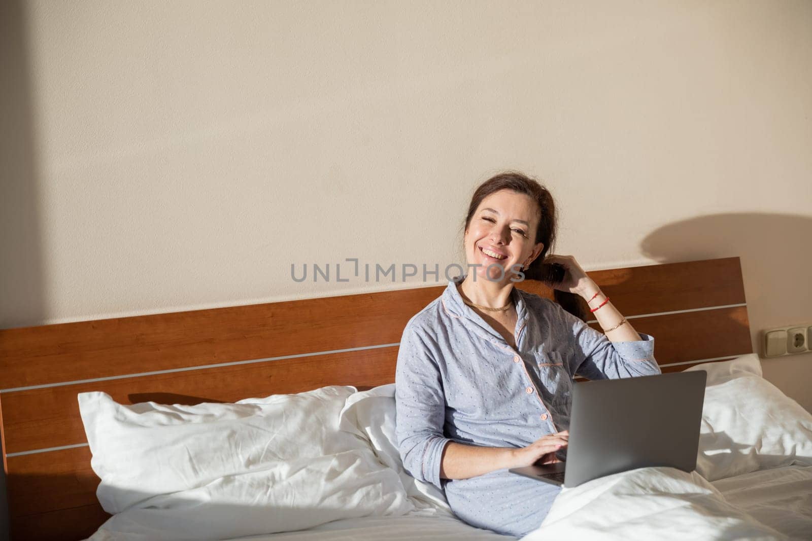 Brunette woman in pajamas with laptop working in bedroom on bed