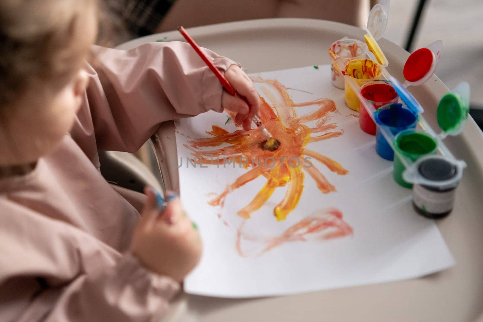 Little girl painting a bright yellow sun with gouache. Childs hand holding a paintbrush, early art skills, imagination and color exploration.