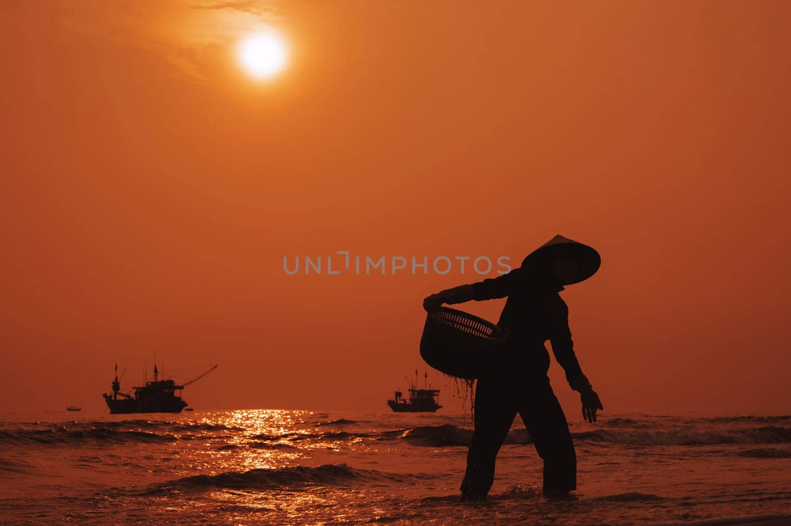 Silhouette of Vietnamese woman in straw hat on beach of sea with ships in fishing market in village in the morning at sunrise in Vietnam