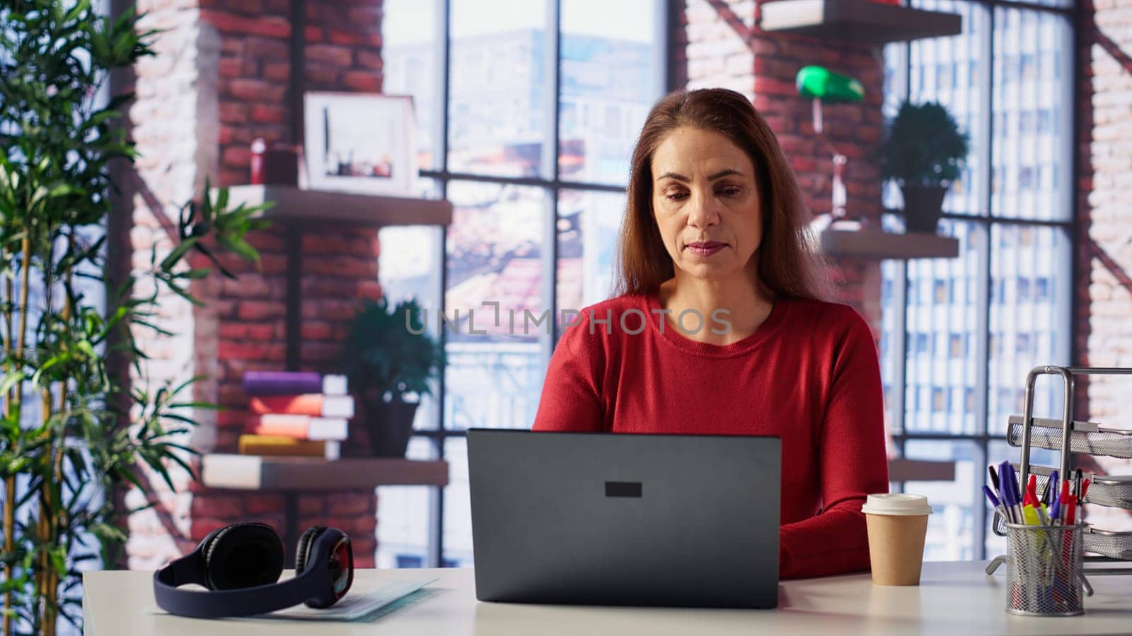 Mature girl freelancer managing her creative projects remotely in loft, working on a new novel or script for her gig. Woman focusing on writing and editing her creation, telework. Camera A.