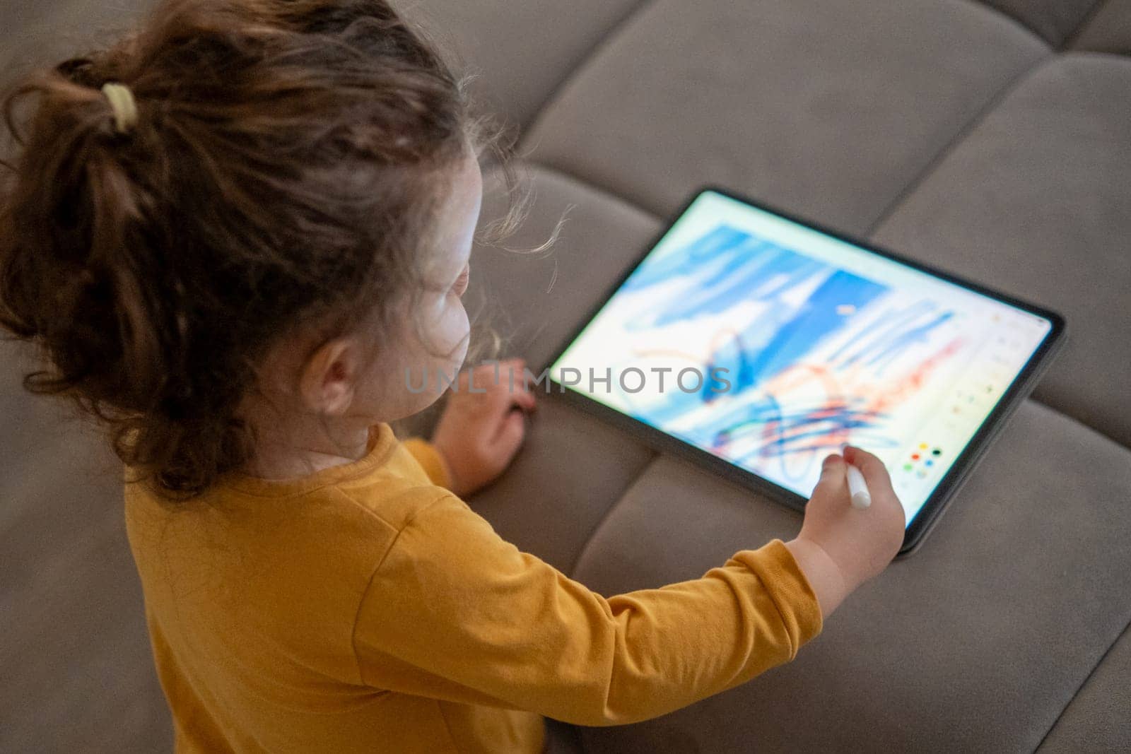 Top view little girl drawing on a digital tablet in a cozy living room. Modern childhood creativity with technology.