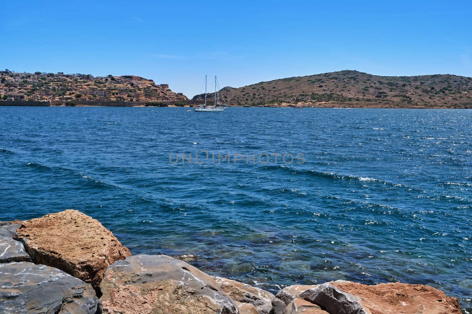 Spinalonga island, Venetian fortress and anchored yacht in lagoon with breakwater stones in foreground on clear sunny summer day, Crete, Greece. film grain