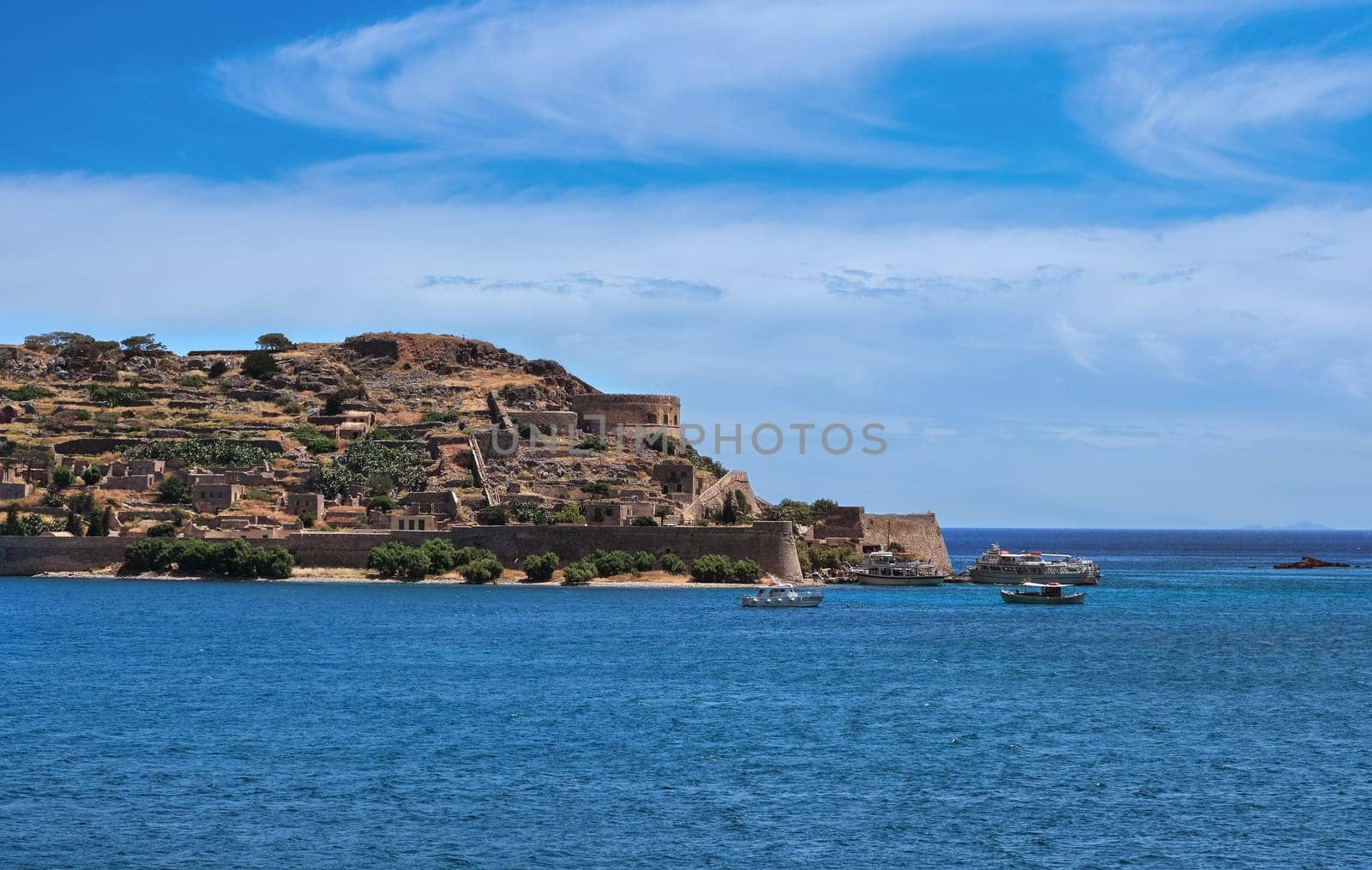 Beautiful view of west bank of Spinalonga island and Venetian fortress on clear sunny summer day on Crete, Greece. Boats passing, old fortifications, calm blue sea. film grain