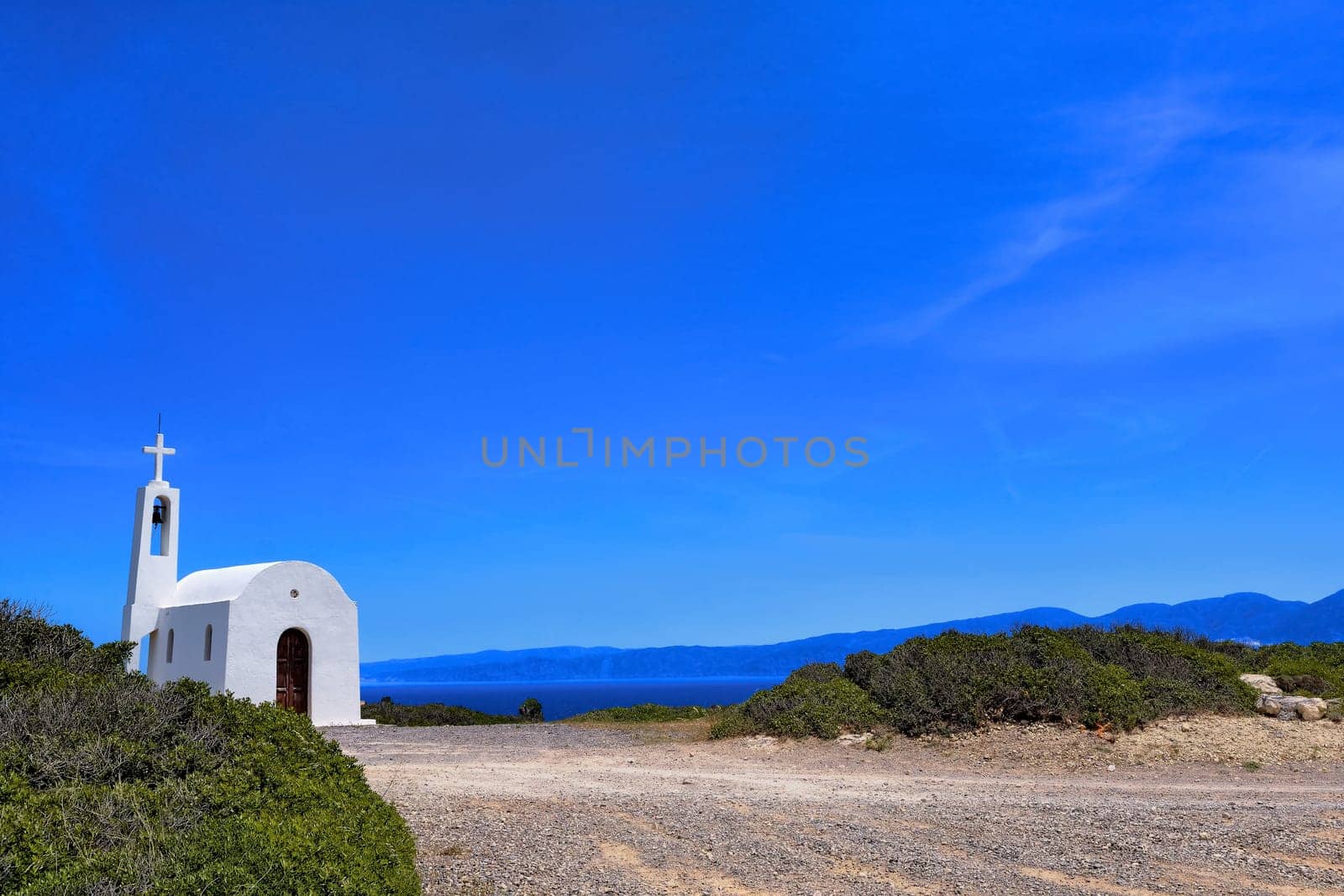 White Greek Orthodox chapel or church on hilltop of seashore and road to it against clear blue sky on sunny day. Traditional religious building. Typical Greek landscape. Diagonal composition