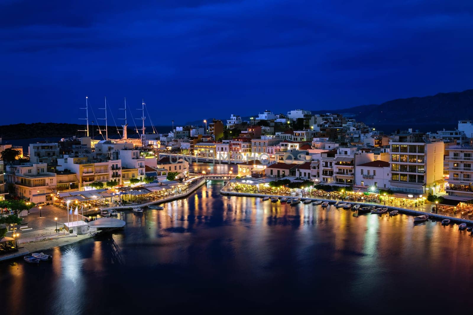 Colorful view of Voulismeni lake and Agios Nikolaos town on Crete island, Greece at dusk. Boats, cafes, restaurants, ship, marina, promenade.
