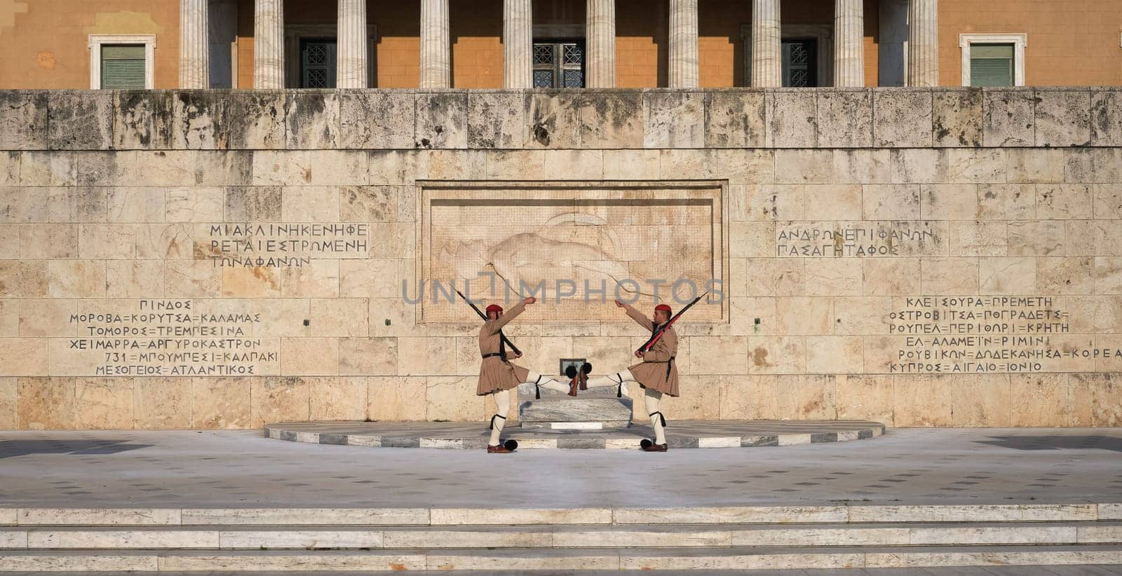 Ceremony of Presidential guard Evzones by the Greek tomb of Unknown Soldier. Evzones in summer uniform face each other, hands up, legs up. Parliament building. Wide shot.