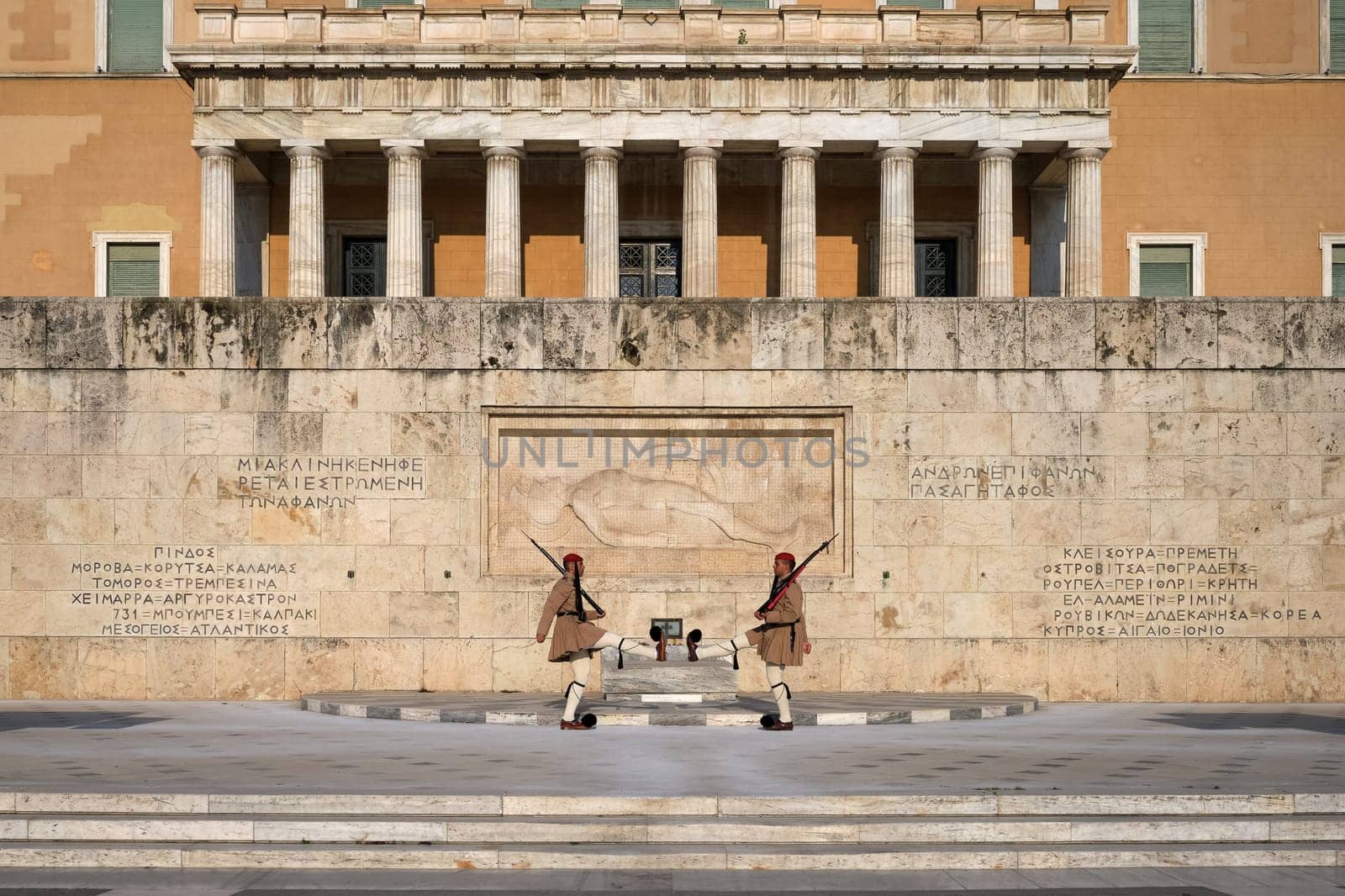 Ceremony of Presidential guard Evzones by the Greek tomb of Unknown Soldier. Evzones in summer uniform face each other, legs up. Parliament building. Wide shot.
