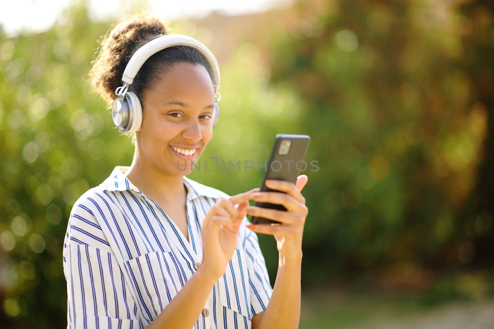Happy black woman wearing headphone and holding smart phone posing looking at camera listening to music in a park