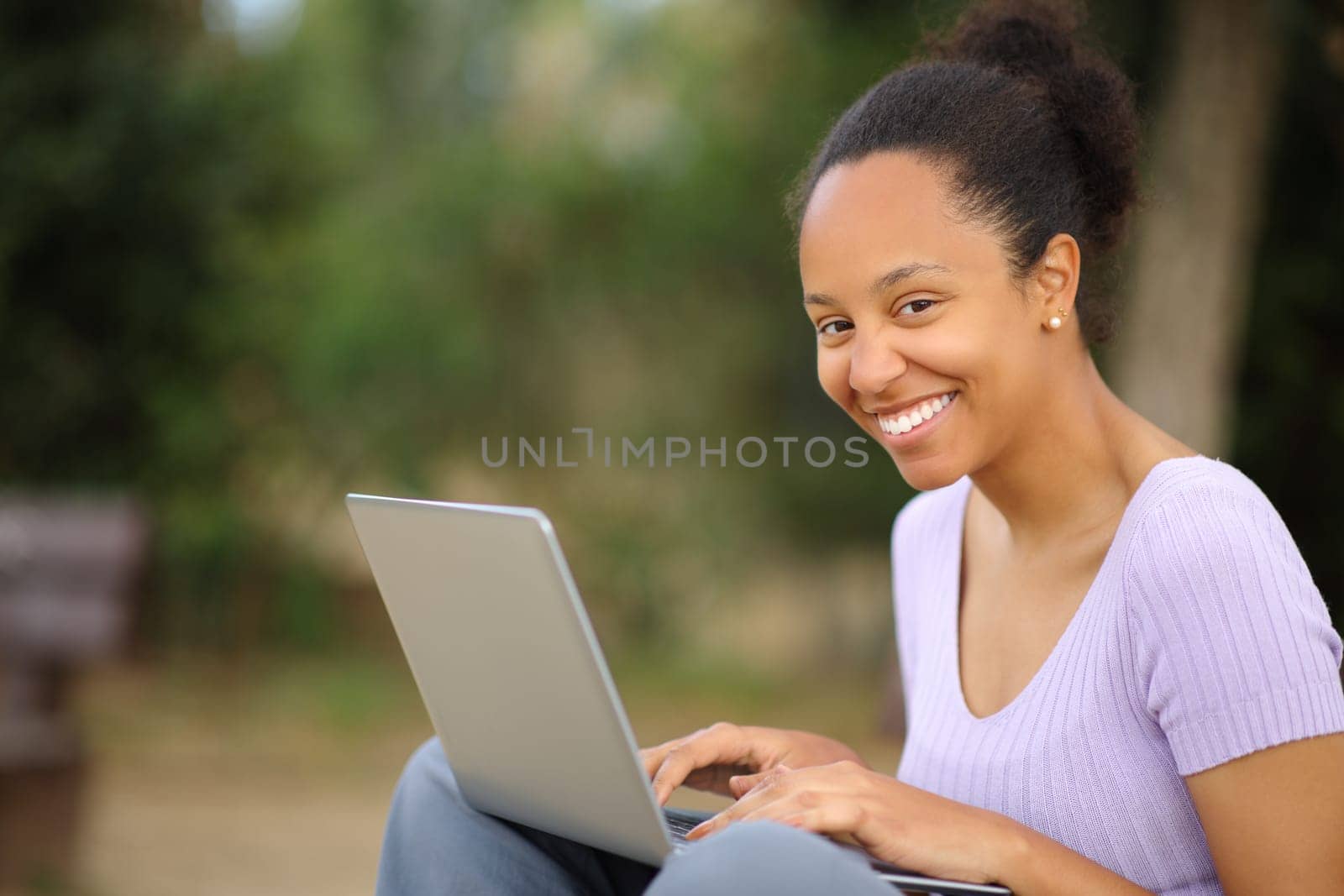 Happy black woman using laptop looking at you sitting in a park